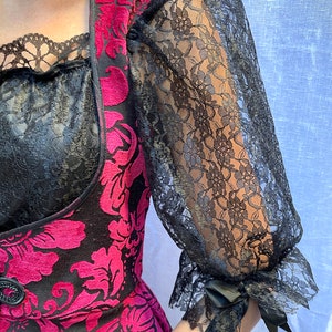 May include: A pink and black velvet dress with a floral pattern. The dress has a square neckline and long sleeves with black lace cuffs. The sleeves are sheer and have a gathered look at the cuff.