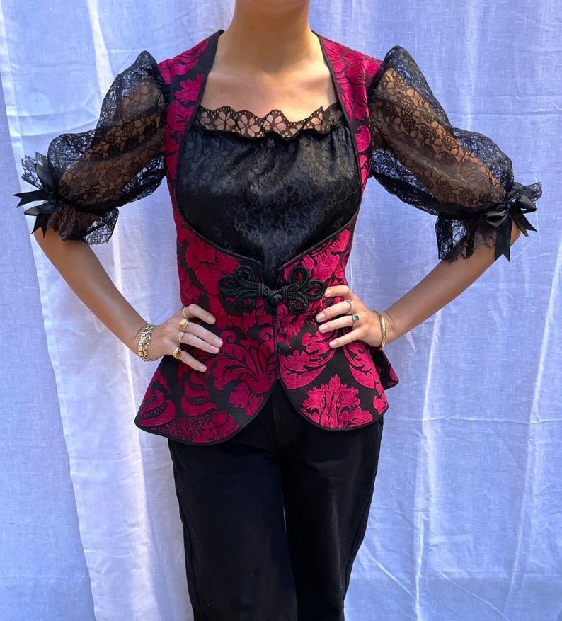 May include: A red and black brocade vest with a black lace trim and a black lace overlay. The vest has a fitted bodice and a peplum hem.