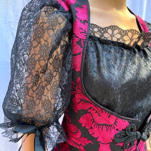 May include: A black and pink velvet vest with a floral pattern and black lace trim. The vest has a black bow at the waist and a black lace overlay on the neckline.