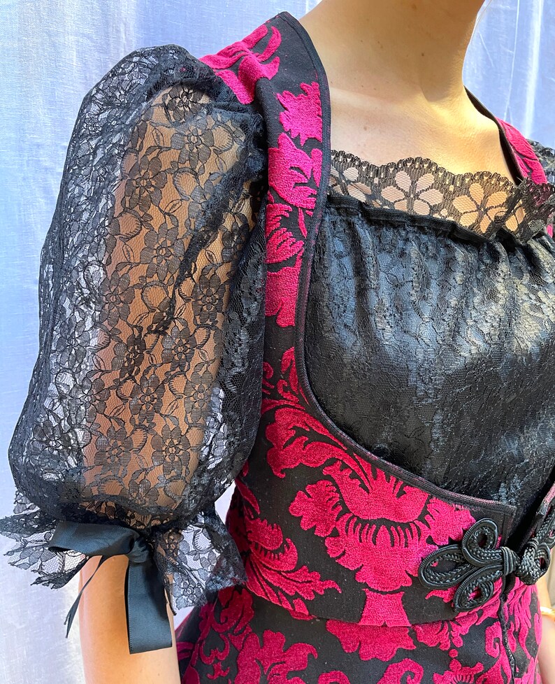 May include: A black and pink velvet vest with a floral pattern and black lace trim. The vest has a black bow at the waist and a black lace overlay on the neckline.
