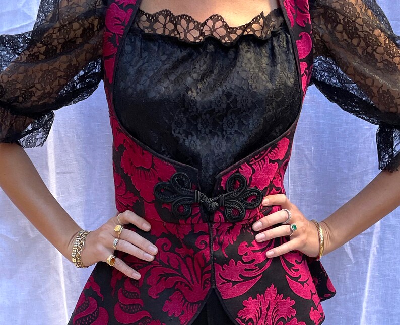 May include: A black and red brocade vest with a black lace trim and a black satin lining. The vest has a floral pattern and a black bow at the center.