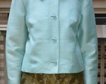 May include: A light blue jacket with a round collar and three decorative buttons. The jacket has long sleeves and a textured appearance. The bottom of the jacket is paired with a green floral patterned skirt. The jacket is a classic style.