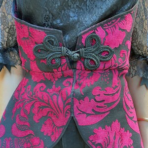 May include: A black and burgundy corset with a floral pattern and a black bow detail. The corset has a lace trim and a fitted silhouette.