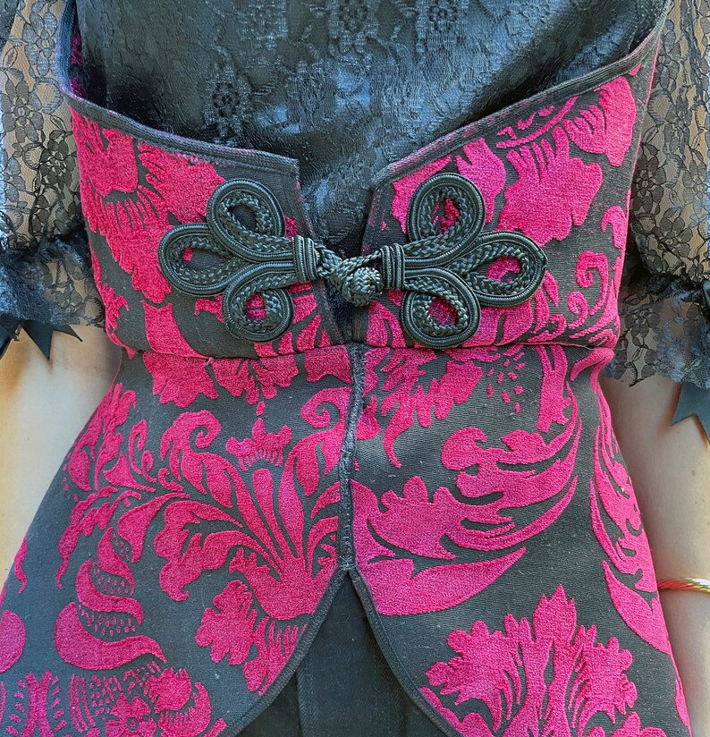 May include: A black and burgundy corset with a floral pattern and a black bow detail. The corset has a lace trim and a fitted silhouette.