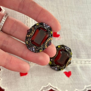 May include: A pair of vintage earrings with a large, dark red, faceted, rectangular stone set in a gold-toned metal frame. The frame is decorated with smaller purple, green, and gold stones.