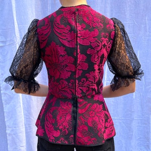 May include: A black and pink patterned blouse with a peplum hem and sheer lace sleeves. The blouse has a fitted bodice and a loose, flowing skirt.