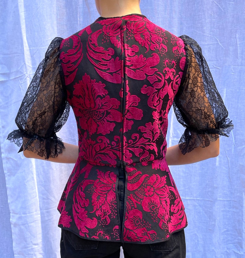 May include: A black and pink patterned blouse with a peplum hem and sheer lace sleeves. The blouse has a fitted bodice and a loose, flowing skirt.