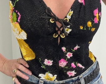 Può includere: Un top in pizzo nero con stampa floreale e una chiusura con lacci sul davanti. Il top ha una scollatura a V ed è indossato con una cintura intrecciata nera.