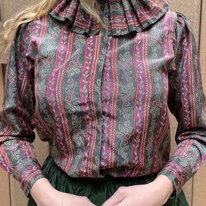 May include: A long-sleeved, button-up shirt with a ruffled collar. The shirt is made of a patterned fabric with vertical stripes in shades of green, pink, and gold. The pattern features small floral motifs.