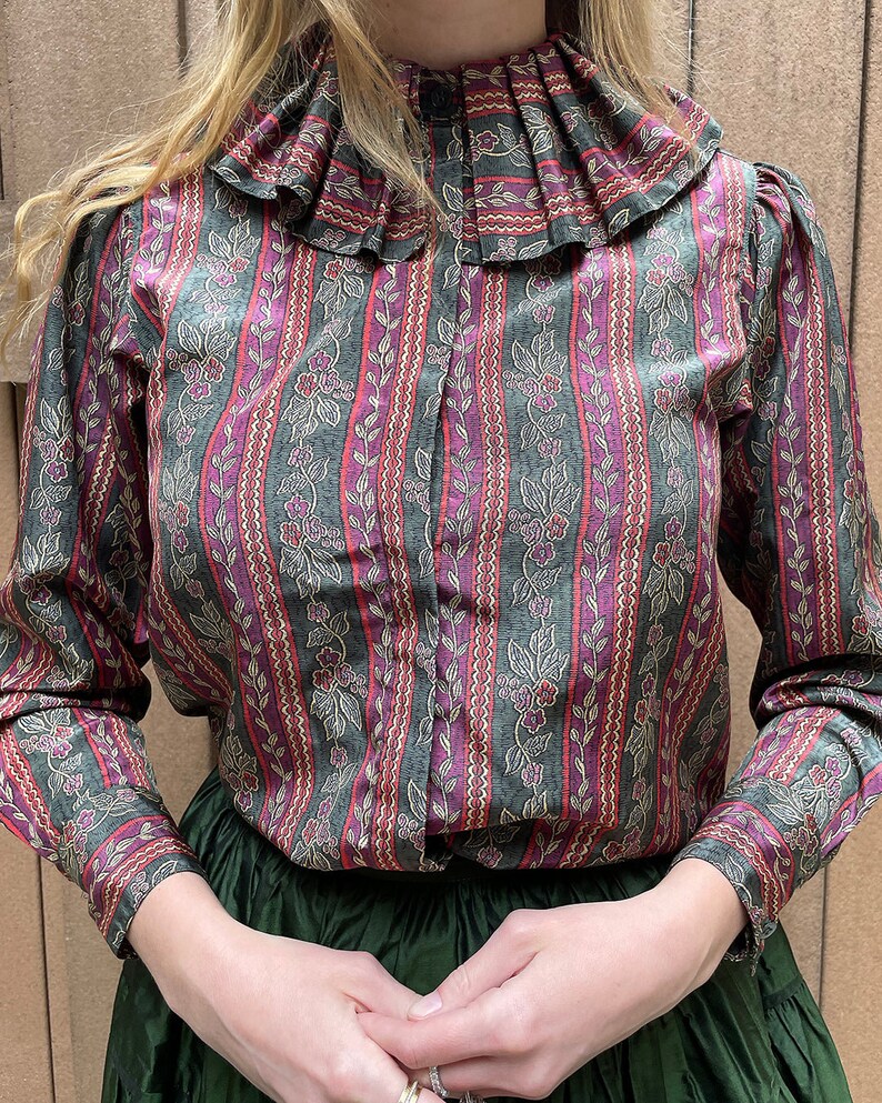 May include: A long-sleeved, button-up shirt with a ruffled collar. The shirt is made of a patterned fabric with vertical stripes in shades of green, pink, and gold. The pattern features small floral motifs.