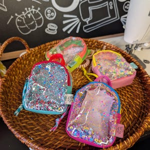 May include: A collection of small, colorful, glitter-filled backpack-shaped pouches with clear fronts, displayed in a woven basket. Each pouch has a different color scheme, including pink, blue, and yellow, with matching zippers and straps.