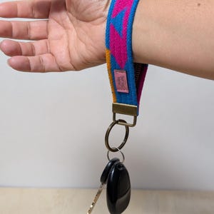 以下が含まれることがあります： ピンク、青、黄色の幾何学模様のカラフルなリストキーフォブ。 キーフォブは、2つの黒い車のキーを保持する金属リングに取り付けられています。 リストストラップには、テキストが記載された小さなピンク色のタグが付いています。