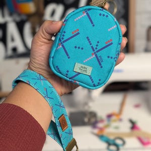 May include: A teal pouch and wrist strap set with a geometric pattern in pink, red, and navy blue. The pouch has a small loop and a keychain ring. The wrist strap has a metal clasp and a brown leather tag. The text "CREATE FUSION" and "PDX" are visible.