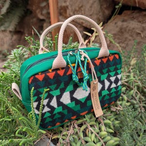 May include: A small, green rectangular handbag with a patterned front featuring black, green, orange, and white geometric shapes. The bag has beige handles and a zipper closure. A small tag that says "Handmade" is attached.