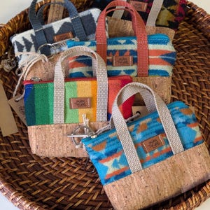 May include: A collection of small, colorful tote bags with cork bases and patterned fabric tops. Each bag features a zipper closure, sturdy handles, and a small leather tag. The bags are displayed in a woven basket.
