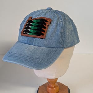 May include: A light blue denim baseball cap with a decorative patch. The patch features a brown border and a colorful design with green, black, and orange elements. The cap is displayed on a mannequin head.