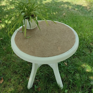 Op de afbeelding: Ronde witte bijzettafel met een geweven juteblad. De tafel heeft drie poten en staat op groen gras.