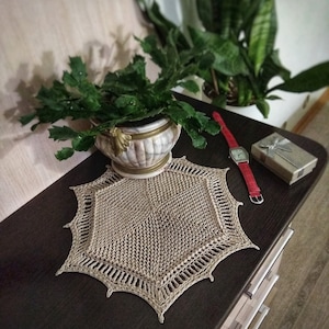 Könnte beinhalten: Eine beige gehäkelte Tischdecke mit sechseckiger Form und einem gewellten Rand. Die Tischdecke liegt auf einer dunkelbraunen Holzoberfläche mit einer Topfpflanze und einer roten Uhr.