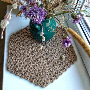 Puede incluir: Un mantel individual de crochet marrón con un patrón de estrella. El mantel individual está sobre una superficie blanca con un jarrón verde y flores moradas.