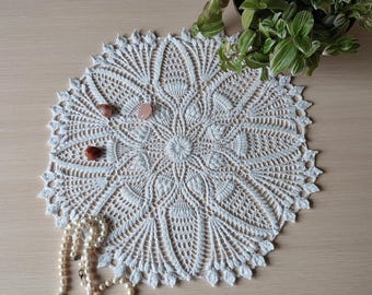 May include: White crocheted doily with a delicate floral pattern. The doily is laid flat on a light wood surface with a strand of pearls and three small pink and orange stones.
