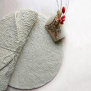 Könnte beinhalten: Ein runder, beiger Häkeluntersetzer mit einem aufgerollten Untersetzer darauf. Der Untersetzer liegt auf einer hellrosa Oberfläche. Eine weiße Kerze mit einem dekorativen Metalldesign und roten Beeren befindet sich in der oberen rechten Ecke des Bildes.