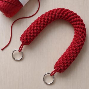 May include: A curved, deep red crocheted handle featuring a thick, textured knot pattern. The handle is finished with a silver-toned metal ring securely attached to each end. A matching ball of red yarn is positioned near the top of the handle.