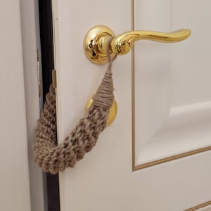 Könnte beinhalten: Nahaufnahme einer Tür mit einem goldenen Türgriff und einem braunen Türstopper aus Seil. Der Türstopper ist aus gewebtem Material gefertigt und am Türgriff befestigt.