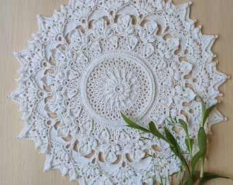 May include: A white crocheted doily with a delicate floral pattern. The doily is circular and has a center with a smaller, more intricate design. The doily is laid on a wooden surface and has green foliage in the foreground.