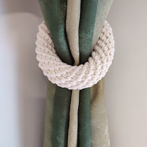 Peut inclure: Un lien de rideau en crochet blanc en forme d'anneau, tenant ensemble deux rideaux à rayures vertes et beiges.