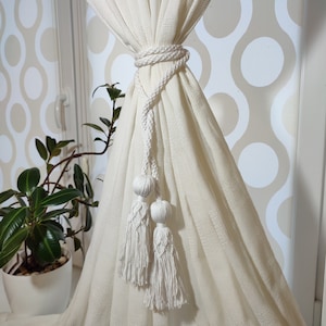 May include: Cream-colored curtain tieback with a braided rope design. The tieback features two decorative tassels and two spherical accents. The curtain is a light cream color, and the background has a retro pattern.