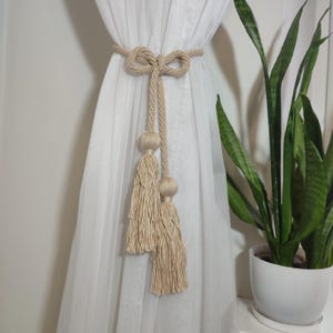 May include: Beige macrame curtain tieback with a knotted bow and two tassels. The tieback is made of a thick, woven material and is used to hold back a sheer white curtain. A potted snake plant is visible in the background.
