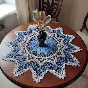 May include: A round wooden table with a blue and white crocheted doily. A small blue vase with gold accents holds three crocheted tulip flowers in beige and yellow. The doily has a starburst design.