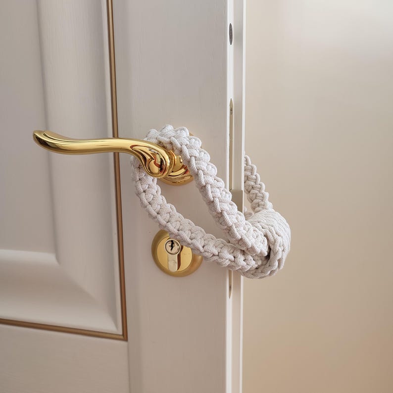 May include: A cream-colored macrame cord loop with a knotted design, wrapped around a polished gold-tone metal door handle. The handle is attached to a white door with a gold-tone keyhole plate featuring a standard cylinder lock mechanism.