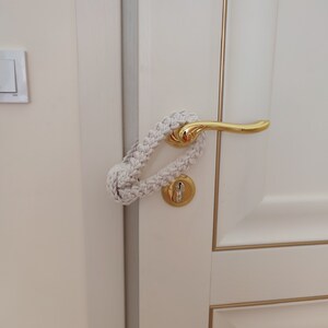 May include: A cream-colored macrame door handle cover featuring a braided cord design and a large knot closure. The accessory is looped around a polished gold-tone metal door handle and keyhole plate on a white door with gold trim accents.