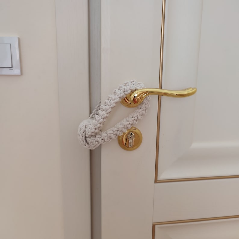 May include: A cream-colored macrame door handle cover featuring a braided cord design and a large knot closure. The accessory is looped around a polished gold-tone metal door handle and keyhole plate on a white door with gold trim accents.