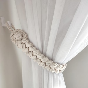 May include: A white cotton curtain with a beige macrame curtain tieback. The tieback is a long, braided rope with a decorative knot at the top.