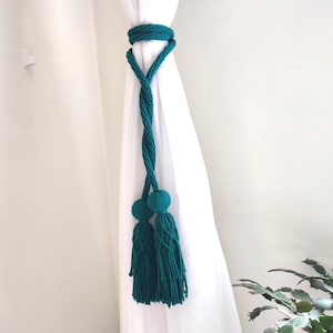 May include: Teal-colored macrame curtain tieback with a twisted rope design. The tieback features two tassels with round ball accents, adding a decorative touch. The tieback is wrapped around a white curtain, creating a stylish home decor accent.