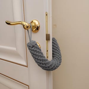 Könnte beinhalten: Ein grauer, gehäkelter Türstopper ist über einer weißen Tür mit einem goldenen Griff angebracht. Der Türstopper besteht aus einem dicken, strukturierten Garn und soll verhindern, dass die Tür zuschlägt. Die Tür und der Türrahmen sind weiß gestrichen.