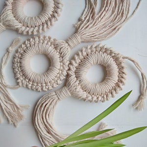 May include: Three beige macrame wall hangings with fringe, each with a circular design. The hangings are on a white surface with green leaves.