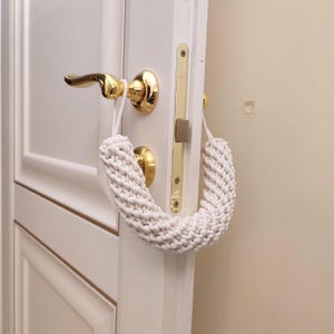 May include: A white macrame door silencer featuring a thick, braided cord design. The accessory is looped over a gold-toned metal door handle and latch mechanism, designed to prevent the door from fully closing and latching shut.
