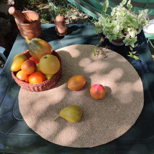 May include: A still life arrangement featuring a round woven placemat with various fruits, including oranges, apples, and a pear. A woven basket holds some of the fruit. A mortar and pestle, a small pitcher, and a potted plant are also present.