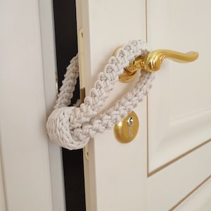 May include: A light gray macrame door handle cover wrapped around a gold-colored door handle. The cover is made of thick, woven cotton rope and is designed to prevent the door from closing. The door is white with gold trim.
