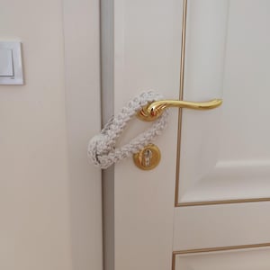 May include: A cream-colored macrame door handle cover featuring a braided rope design with a knotted loop end. The textile accessory is looped securely around a polished gold-tone metal lever handle on a white door with gold trim accents.