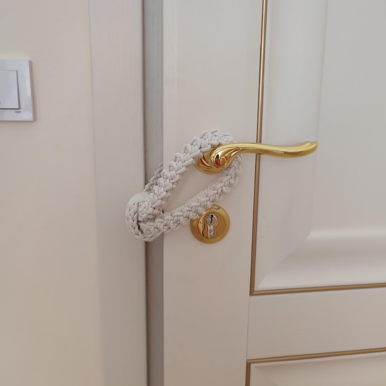 May include: A cream-colored macrame door handle cover featuring a braided rope design with a knotted loop end. The textile accessory is looped securely around a polished gold-tone metal lever handle on a white door with gold trim accents.