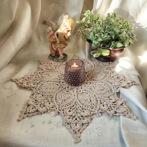 May include: A beige crocheted doily with a delicate floral pattern, centered on a white fabric surface. A small figurine of a fairy with wings and a candle are placed on the doily.