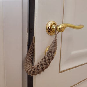 May include: A close-up of a white door with a gold door handle. A brown knitted doorstop is hanging from the handle.