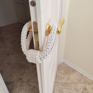 May include: A cream-colored, braided macrame door handle cover featuring a thick, knotted design. The textile accessory is looped around a polished gold-tone metal door handle attached to a white door with a metallic latch plate and hardware.