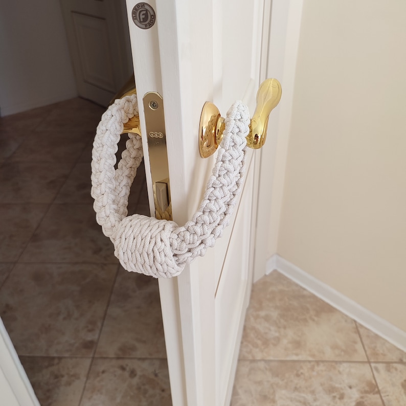 May include: A cream-colored, braided macrame door handle cover featuring a thick, knotted design. The textile accessory is looped around a polished gold-tone metal door handle attached to a white door with a metallic latch plate and hardware.