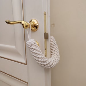 以下が含まれることがあります： 金色のドアノブに、馬蹄形の白い編み物のドアストッパーが掛かっています。ドアストッパーは厚手のざっくりとした糸でできています。