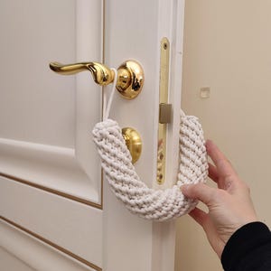May include: A white crocheted doorstop in the shape of a horseshoe hangs on a gold door handle. The doorstop is made of thick, tightly woven yarn.